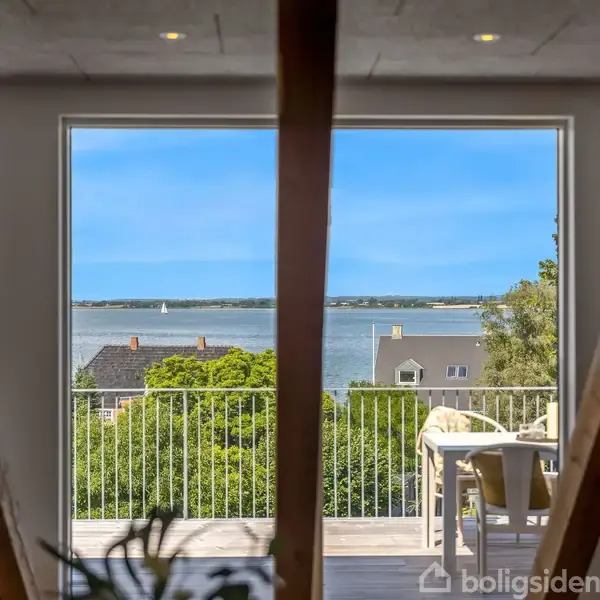Tagterrasse med glasbord og stole placeret på en bygning. Bagved er et skråt tegltag med ovenlysvinduer. Omkringliggende huse har lignende tegltage. Himlen er skyfri og blå.