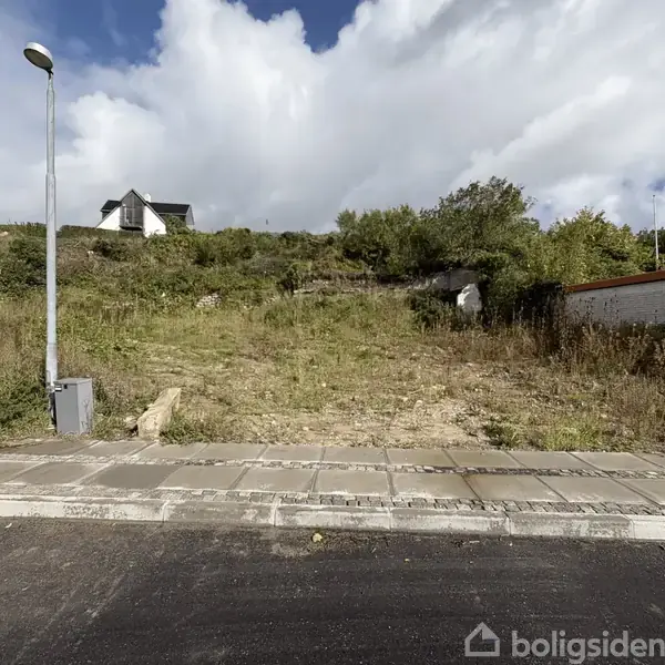 En ubebygget grund med græs og buske ligger op ad en bakket skråning. En enkelt gadelampe står ved siden af fortovet. En hvid bygning ses i baggrunden.