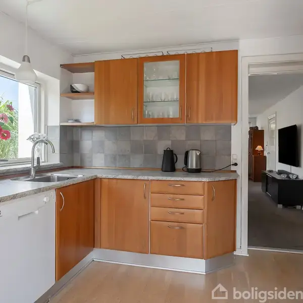 Køkken med træskabe og grå flisevæg indeholder en vask, elkedel og toaster. En vase med blomster står ved vinduet. Døråbning fører til stue med TV på væggen.