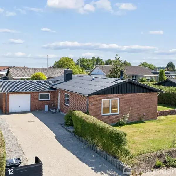 Rødt murstenshus med garage står på en velplejet grund omkranset af hække. Bilindkørsel fører op til huset. Tætliggende huse ses i de omkringliggende kvarterer. Nummer "12" er synligt ved indkørslen.