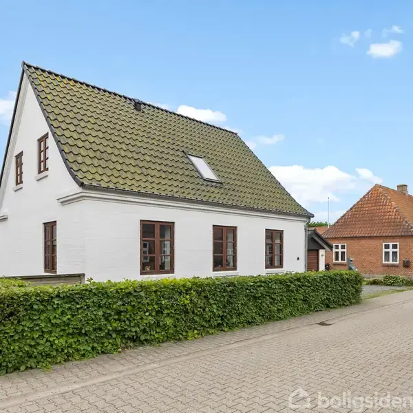 Hvidt hus med grøn tag, står stille, omgivet af en frodig hæk; ved siden af en brolagt vej og et rødt murstenshus.