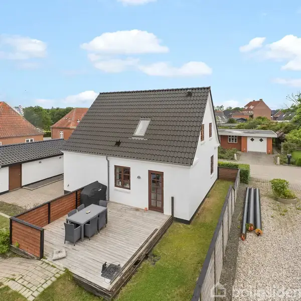 Et hvidt hus med gråt tag står i en baghave. Ved siden af er en træterrasse med havemøbler. Omkring er andre huse og en grusvej.