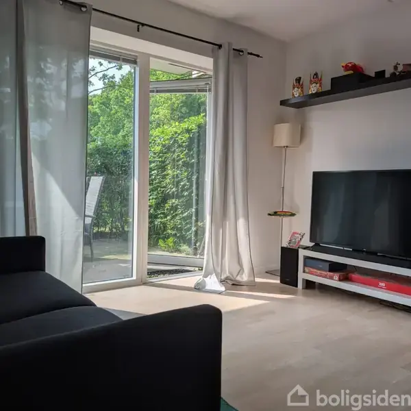 En stue viser en sofa med udsigt til en stor fladskærms-TV på et lavt bord. Store glasdøre fører til en have. Gardiner skaber en lys, venlig atmosfære.