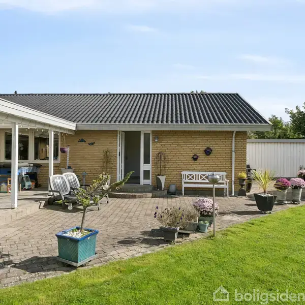 Et gult murstenshus med sort tag har en terrasse med havemøbler og blomsterkrukker. Terrassen fører til en grøn græsplæne omkranset af buske og træer.