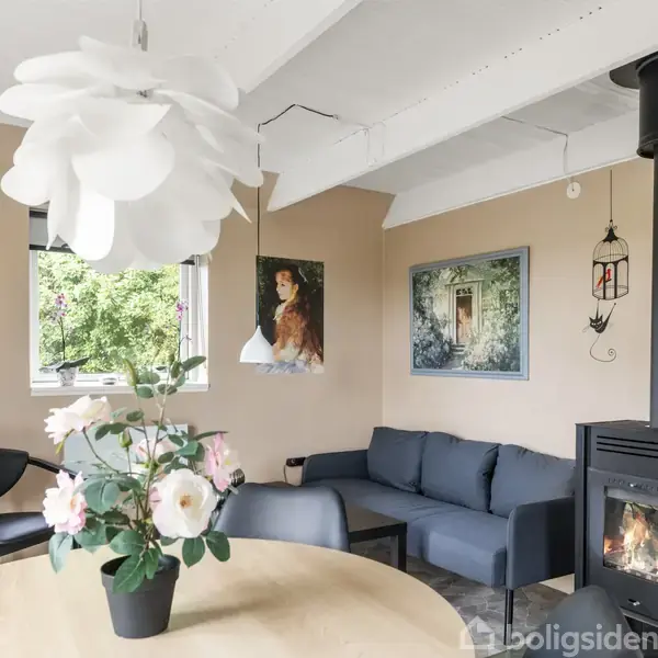 En hyggelig stue med en blå sofa står ved siden af en sort brændeovn. En rund spisebord med en vase med blomster står foran. Kunstværker hænger på de lyse vægge.