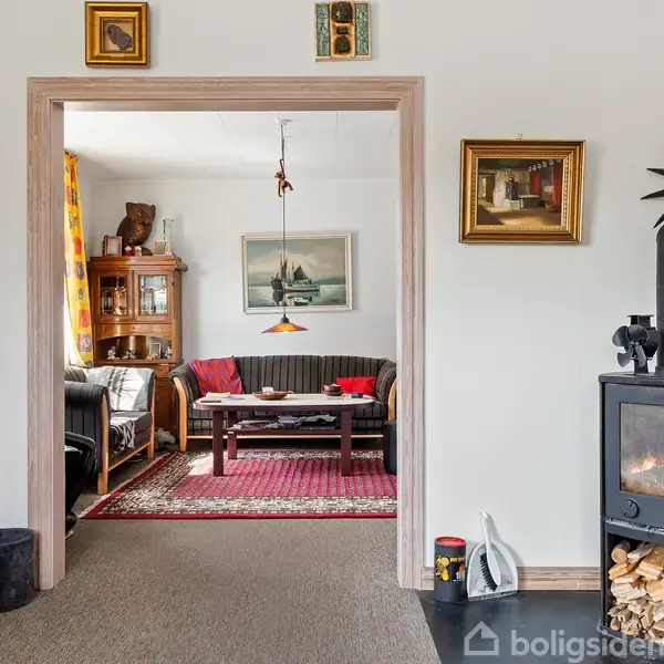 Brændeovn brænder i stue med vægge dekoreret med malerier. Åbning fører til rum med sofa, bord og tæppe. Træ er stablet ved ovnen.