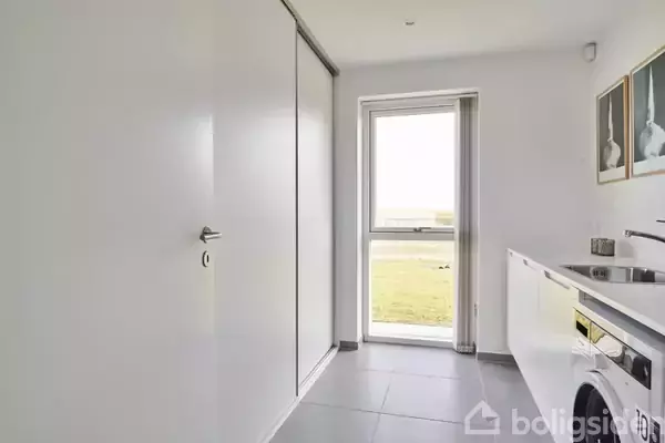 Vaskemaskine står i lyst vaskerum med gråt flisegulv, ved siden af en hvid arbejdsbænk. Der er store skabe til venstre og en glasdør fører ud til en græsplæne.