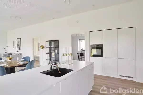 Et moderne køkken med hvidt interiør har en stor kogeø og integreret ovn. I baggrunden er et spisebord med blå stole og dekorative elementer i et lyst rum.