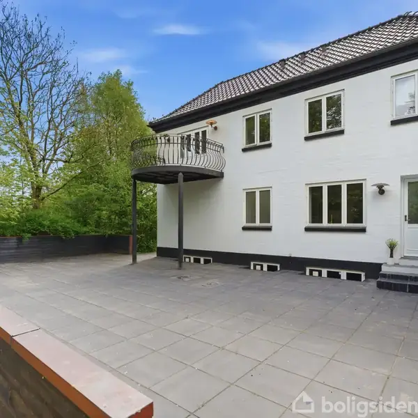 To-etagers hvidt hus med en balkon vender mod en stor terrasse med fliser. Omgivet af grønne træer under en klar blå himmel.
