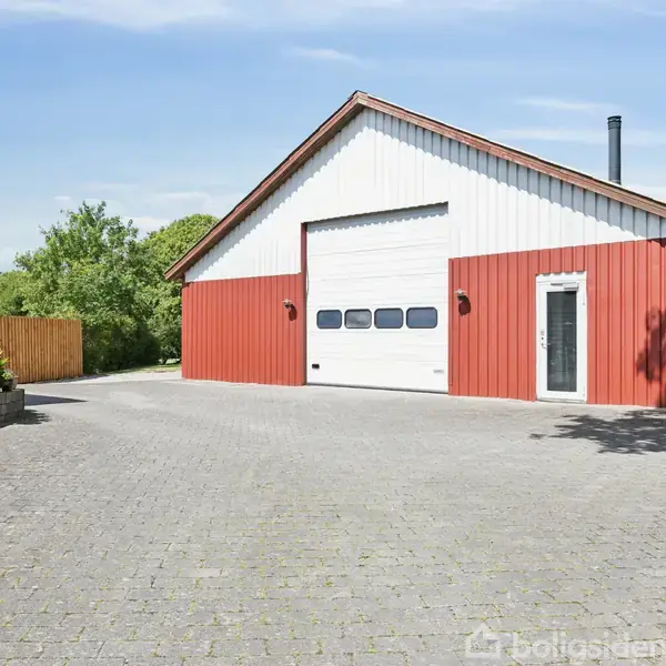 En stor rød garagebygning med hvid port ses. Den står på en brolagt gårdsplads omgivet af grønt løv og træer på en solrig dag.