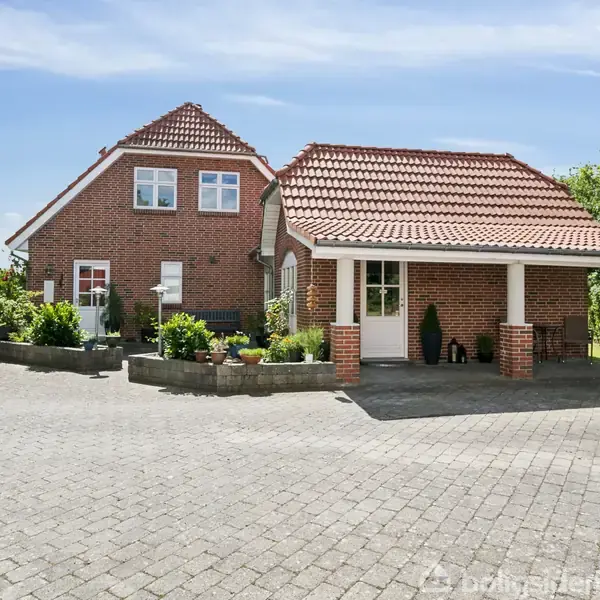 Rødstenshus står stille på en flisebelagt indkørsel, omkranset af frodige planter og grønne træer. Huset har rødt tegltag og hvide vinduer, omkranset af en klar blå himmel.