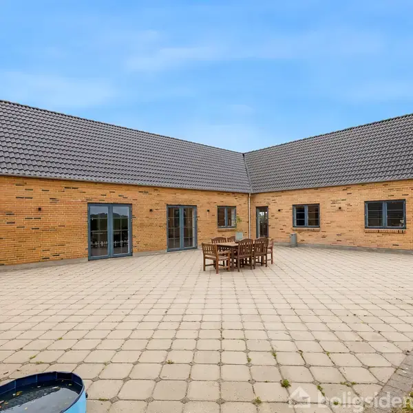 En L-formet murstensvilla står på en stor flisebelagt terrasse med et havebord og stole i centrum, omgivet af en grøn græsplæne og under en klar blå himmel.