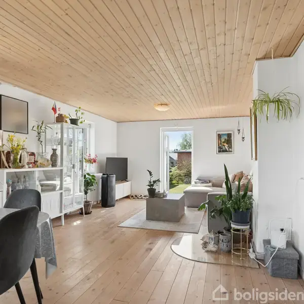 Stue med træloft og trægulv med spisebord og stole til venstre. Reoler med bøger og dekorationer langs væggen. Sofa og sofabord nær glasdør med udsigt til have.