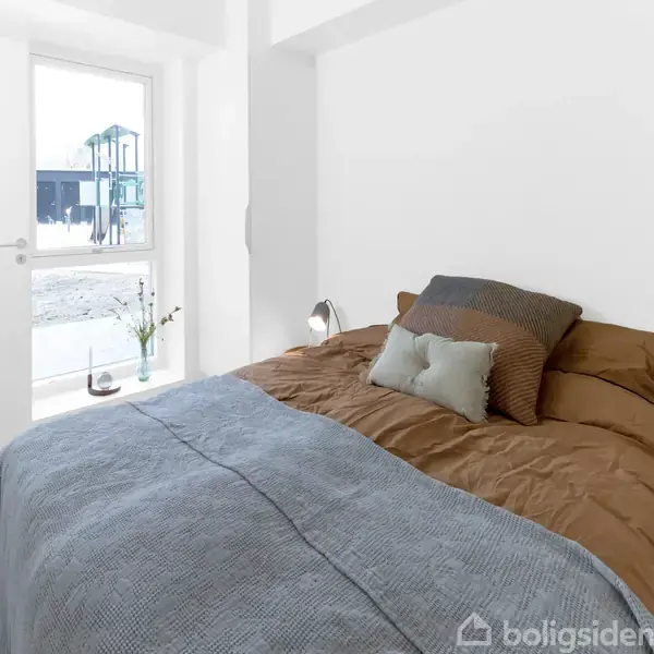 Seng med brun og grå dyne, pyntepuder. Stående ved siden af er en lampe. Lysindfald fra vinduet, vask i vaser på vindueskarmen. Hvidmalet værelse med trægulv.