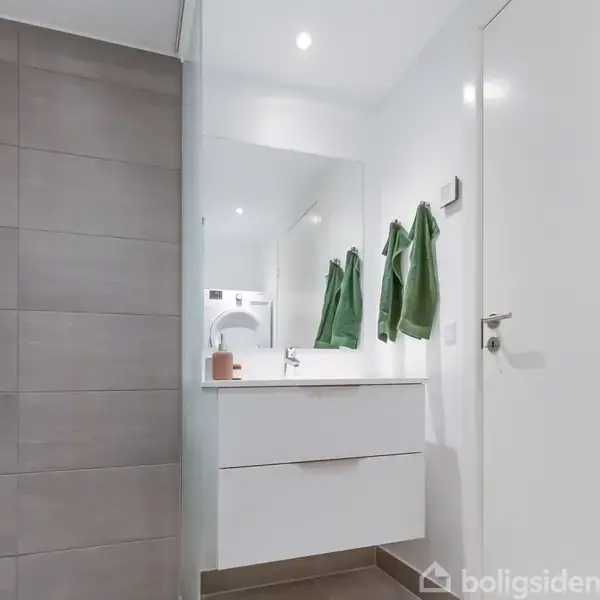 Badeværelse med hvidt skabsmodul under vask, spejl på væggen. Grønne håndklæder hænger på kroge. Grå fliser i bruseniche. Minimalistisk og ren indretning.