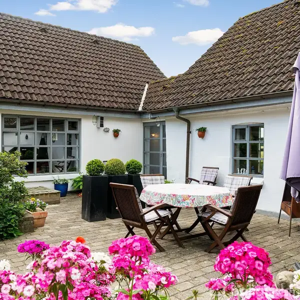 En gårdhave med et rundt bord dækket af dug og fire stole omkring det. Omkring bordet er farverige blomster og grønne planter. Hvide vægge og brune tegltage omgiver gårdhaven.