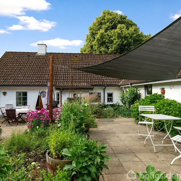En gårdhave med grønne planter og blomster, hvor der står et hvidt bord og stole. Baggrunden viser et traditionelt hus med rødt tag, omgivet af træer og blå himmel.