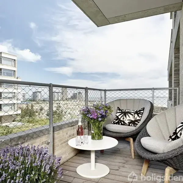 To stole står på en altan med et rundt bord mellem dem. Bordet har blomster og en flaske rosévin. Baggrunden viser moderne bygninger og en blå himmel.