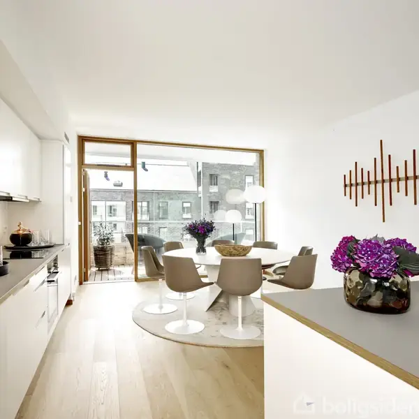 Køkken-alrum med hvide skabe og en træbordplade. Rundt spisebord med fire stole er centralt placeret ved glasdøre til en balkon. Lilla blomster og kunstværk på væggen.