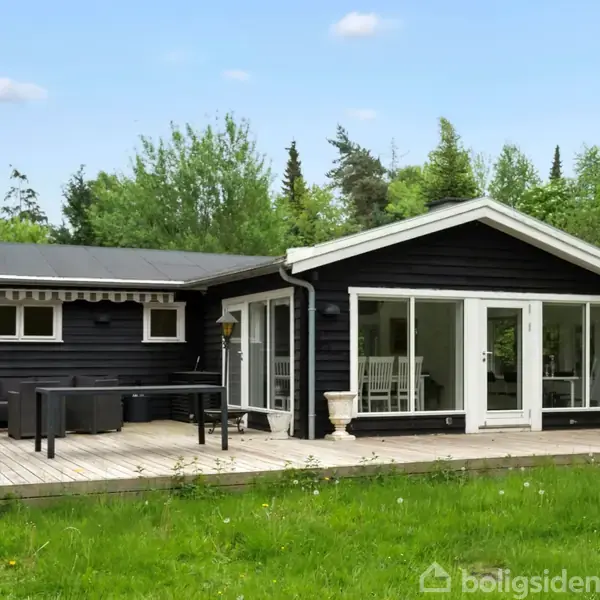 Sort træhus med store hvide glaspartier står på en træterrasse. Terrasseborde og -stole står udenfor, omgivet af grønne træer og græsplæne under en klar, blå himmel.