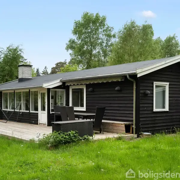 Et sort træhus med hvide vinduesrammer står på en græsplæne, omgivet af grønne træer. Foran huset er der en terrasse med havemøbler.
