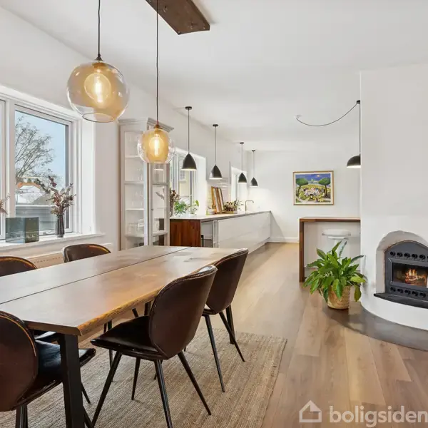 Et langt træbord med seks stole står i en spisestue med trægulv. Store vinduer til venstre lader naturligt lys ind. Rund pejs og grøn plante til højre.