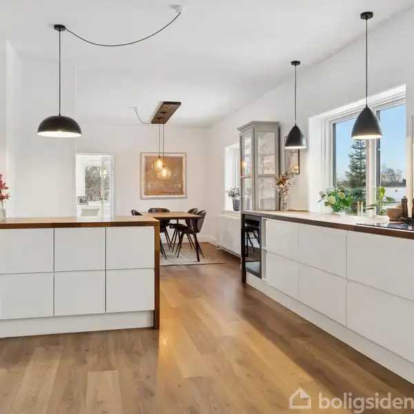 Et moderne køkken har hvide skabe og træbordplader. Tre pendellamper hænger over en central ø. Store vinduer lader sollys komme ind. En forrammet spisestue med bord og stole ses i baggrunden.