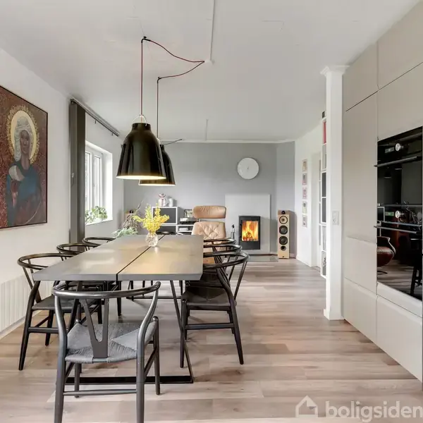 Spisebord med sorte stole, placeret under en stor, hængende lampe, i et moderne køkken-alrum. I baggrunden ses en brændeovn og en lænestol. Vægkunst hænger til venstre.