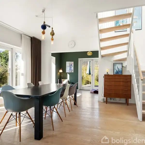 Et moderne spisestueområde med sort bord og seks blå stole står ved store vinduespartier, der åbner op mod en lys have. En trappe fører op til øverste etage.