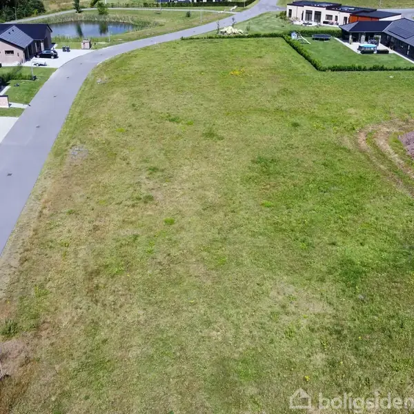 Et grønt græsareal ligger mellem en asfalteret vej og parcelhuse i et bebygget boligområde. Husene har haver, og området er rydeligt og velplejet.