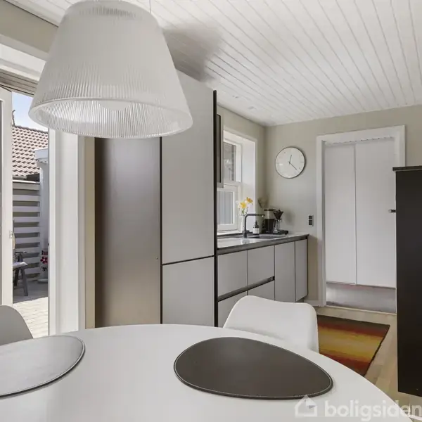 Rundt spisebord med dækkeservietter i en moderne, lys spisestue. En stor lampe hænger over bordet. Køkkenskabe ses i baggrunden sammen med et vindue og en ur på væggen.