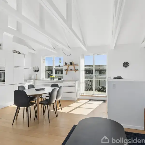 Et lyst køkken-alrum med spisebord, omgivet af hvide skabe og åbenhed. Lys strømmer ind fra store vinduer, der vender ud mod en balkon. En sofa står ved siden af spiseområdet.