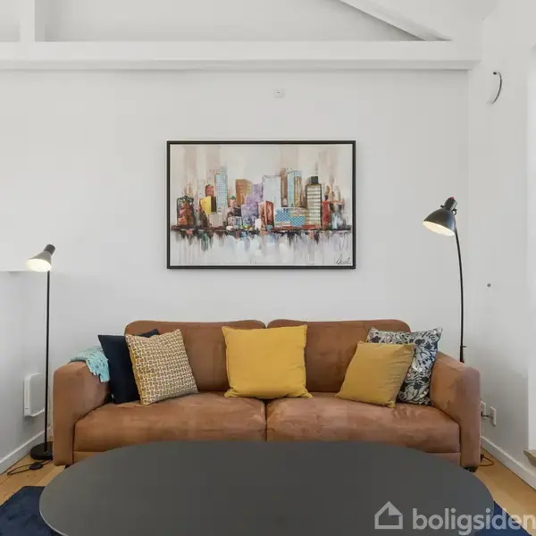 En brun sofa med farverige puder står foran en hvid væg. Over sofaen hænger et maleri af en by. To sorte gulvlamper står på hver side. Ved siden ses et stort vindue.