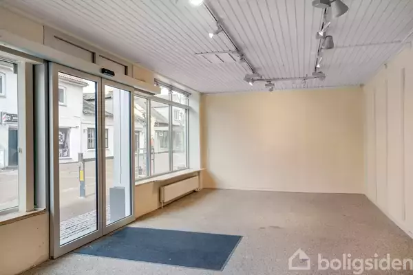 Et tomt butikslokale med glasdør og store vinduer vender ud mod en gade. Indvendigt er der et lyst gulvtæppe og hvidmalede vægge. Loftet har spotlys og en radiator under vinduerne.