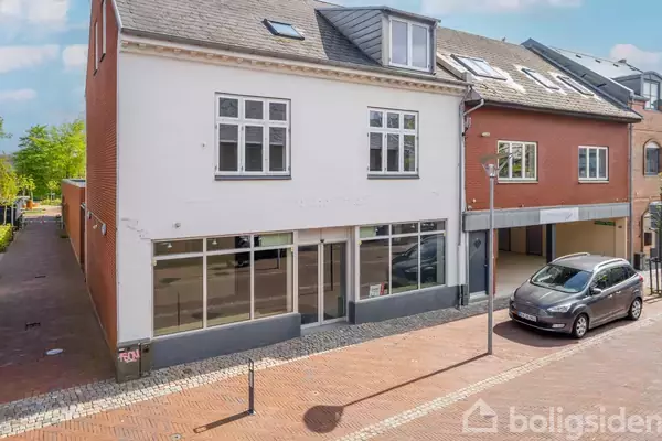 Bygningen er to-etagers med hvid facade og store vinduer. Bil parkeret ved fortovet foran, på en brostensbelagt gade. Baggrund viser blå himmel og nogle træer.