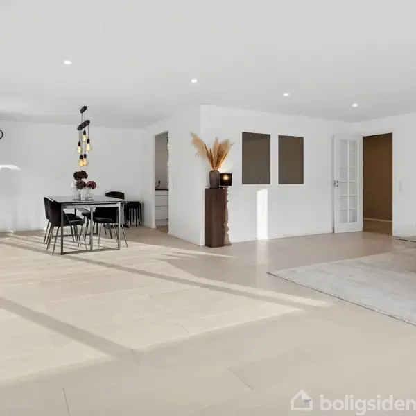 Spisestue med et sort spisebord og stole. Lampe hænger over bordet. Til højre er en stue med lys sofa og tæppe på gulvet. Lyse vægge og minimalistisk indretning.