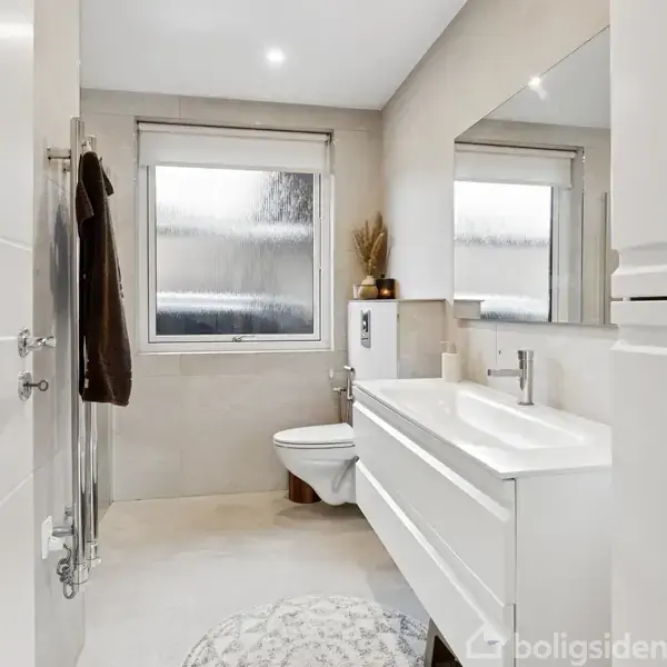 Hvidt badeværelse med væghængt toilet, rummelig vask med skuffer og et stort spejl. Et vindue med matteret glas giver lysindfald, og der hænger et mørkt håndklæde ved bruseren.