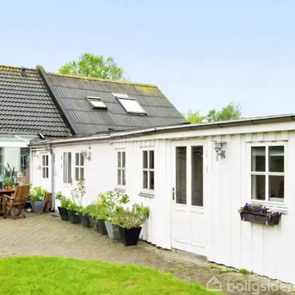 Hvidt hus med sorte tagsten, placeret i grøn have. Et udendørs terrasseområde har en glasveranda og træmøbler. Blomsterkrukker dekorerer stien langs husets væg.