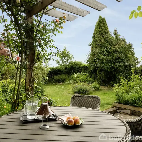 Et rundt bord med træmøbler står på en terrasse. På bordet er en kande vand, glas og en skål med frugt. Omkring ses en frodig have med blomster og buske.