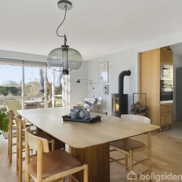 Et spisebord med stole er centralt placeret i en lys spisestue. En stor lampe hænger over bordet. Bagved åbner store vinduer op til en grøn have. En pejs står i hjørnet tæt på køkkenområdet.