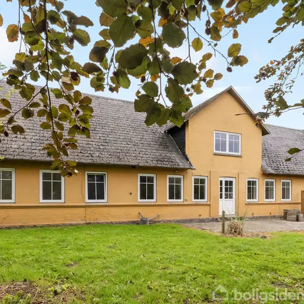 Et gult hus med mørkt tag står i en have med grøn græsplæne og omkransende træer. Huset har flere vinduer og en hvid hoveddør.