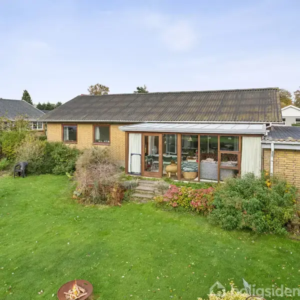 Hus med fladt tag og vinduesparti set fra en grøn have med græsplæne og buske. I baggrunden ses en blå himmel og nabohusenes tage.