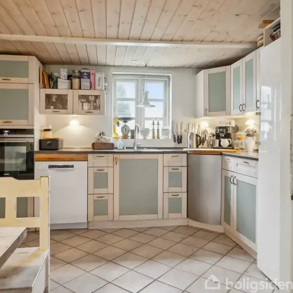 Et køkken med hvide skabe og glaslåger står langs væggen. En ovn er indbygget ved siden af et lille vindue. Træborde fylder rummet med en lys, åben atmosfære.