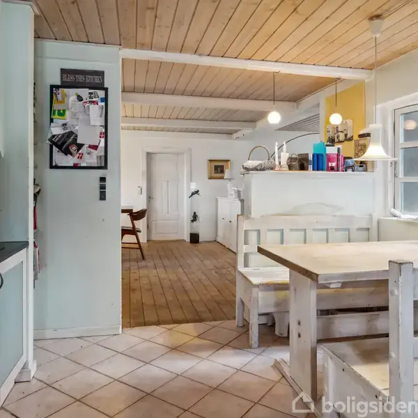 Køkken med trækøkkenbord og bænke placeret i et lyst rum med træloft. Køleskab og opslagstavle på venstre væg, åbning fører til spiseområde med stole i baggrunden.