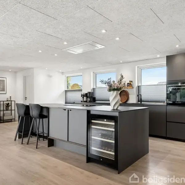Køkken i moderne stil med køkkenø, hvorpå der står en vase med blomster. Tre barstole står ved øen. I baggrunden ses lyse træpaneler og indbyggede skabe med ovn.