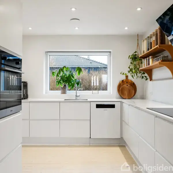 Hvidt køkken med indbyggede apparater og en vask under et vindue, der vender ud mod en have. Hylder med krydderier og planter tilføjer en hjemlig atmosfære.