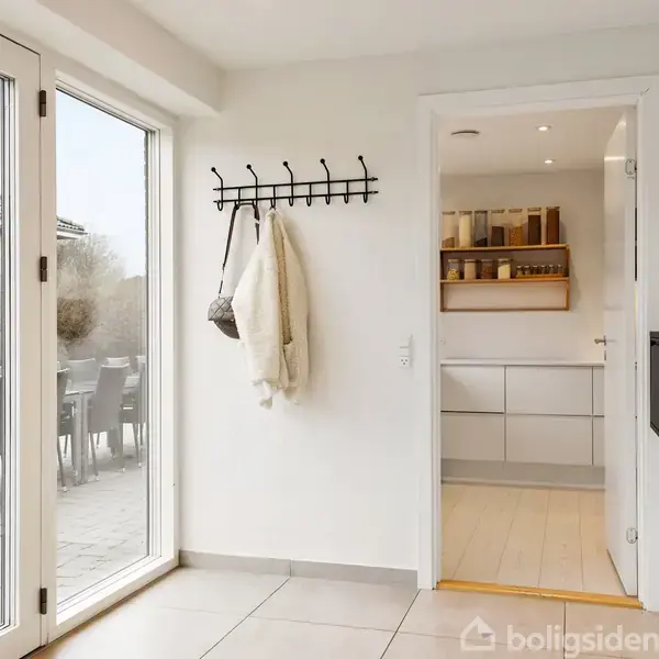 En entré med en knagerække med jakker, glasdøre fører til terrasse med udendørs spisebord og stole, indgang til køkken med hylder og skabe.