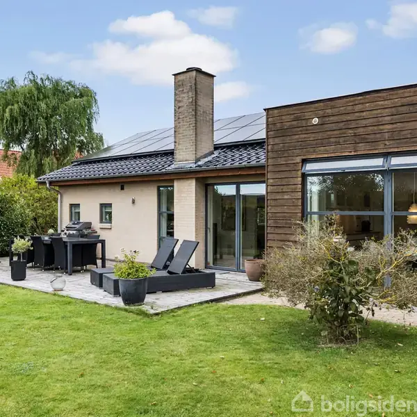 En moderne villa med store vinduer og solpaneler på taget står i en velplejet have med græsplæne. På terrassen er der solsenge og en grill ved siden af dekorative buske.