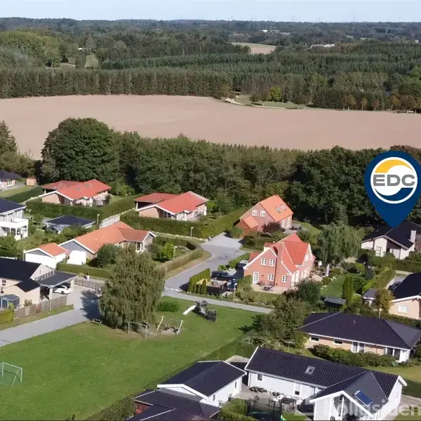 Boligområde med parcelhuse og haver ligger omringet af grønne marker og skove. En markør med teksten "EDC" viser en specifik ejendom blandt de mange huse i landskabet.
