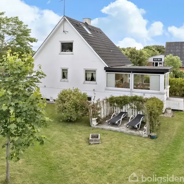 Et hvidt hus med en mørk tag, to etager, store vinduer og en veranda i en velholdt have med grøn græsplæne, to liggestole og omkringliggende buske og træer.